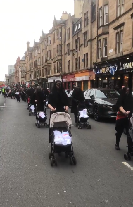 Women dressed in black, faces covered with black veils, push strollers with white bundles in them at the head of the march
