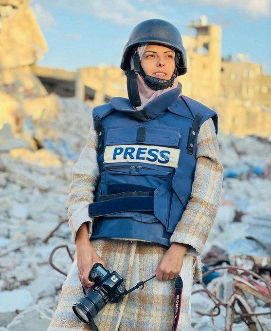 A woman in a blue helmet and press vest standing in the rubble in Gaza, holding a camera