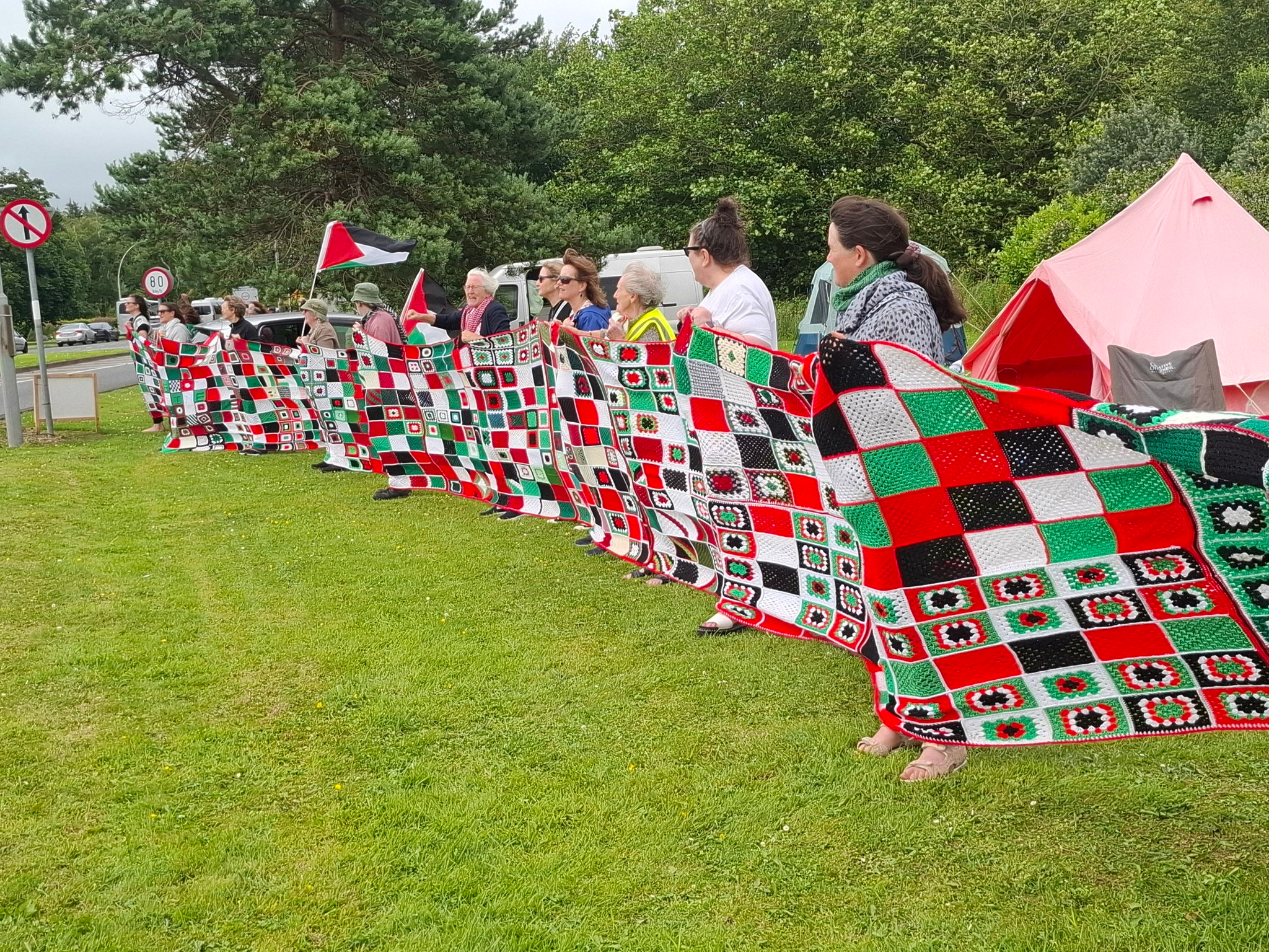 Picture of a line ofpeople holding the peace blanket, a long crocheted blanket where each square represents 30 dead Palestinians, outside Shannon Airport