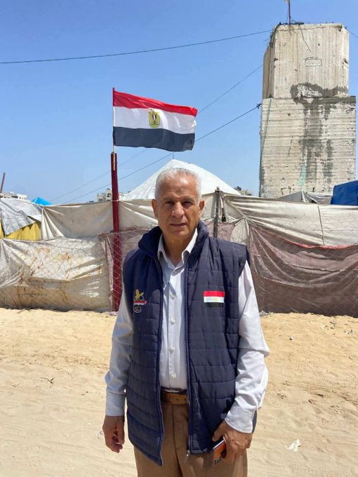 A middle aged man in khaki trousers, a pale shire and a dark blue padded vest jacket, standing in front of some tents.  The Egyptian flag is flying in the background.