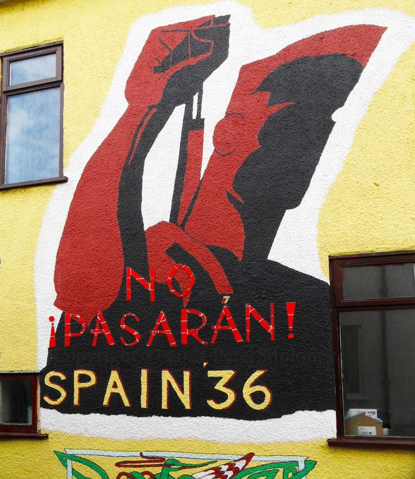 Image of the wall of a house painted bright yellow.  Centre is a red and black stylised image of a fighter with a rifle on their back and fist raised.  In large letters beneath:

¡NO PASARÁN!
SPAIN 36